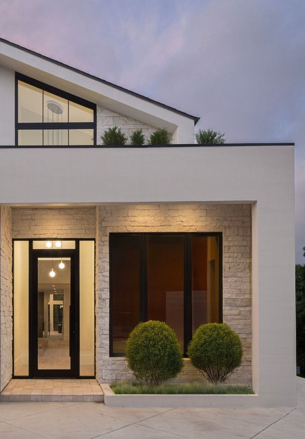 Front exterior view of a refined two-storey residence at dusk. The facade combines clean white ribbed cladding, pale stacked-stone wall sections, dark-framed glazing, a projecting upper volume, and a sheltered entry with warm interior lighting. Neatly clipped shrubs and a concrete forecourt complete the contemporary composition.