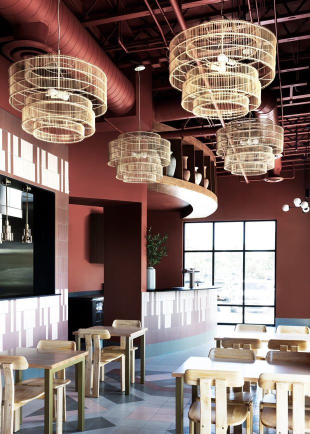 A richly styled commercial dining and hospitality interior, likely a restaurant or cafe. The space features deep terracotta walls, an exposed dark ceiling with large ductwork, multiple oversized tiered wire pendant chandeliers, a curved mezzanine or balcony edge, tiled counter fronts, black-framed glazing, and numerous light timber tables and chairs. Geometric floor tiles in muted pink, blue, grey, and cream create a patterned floor.