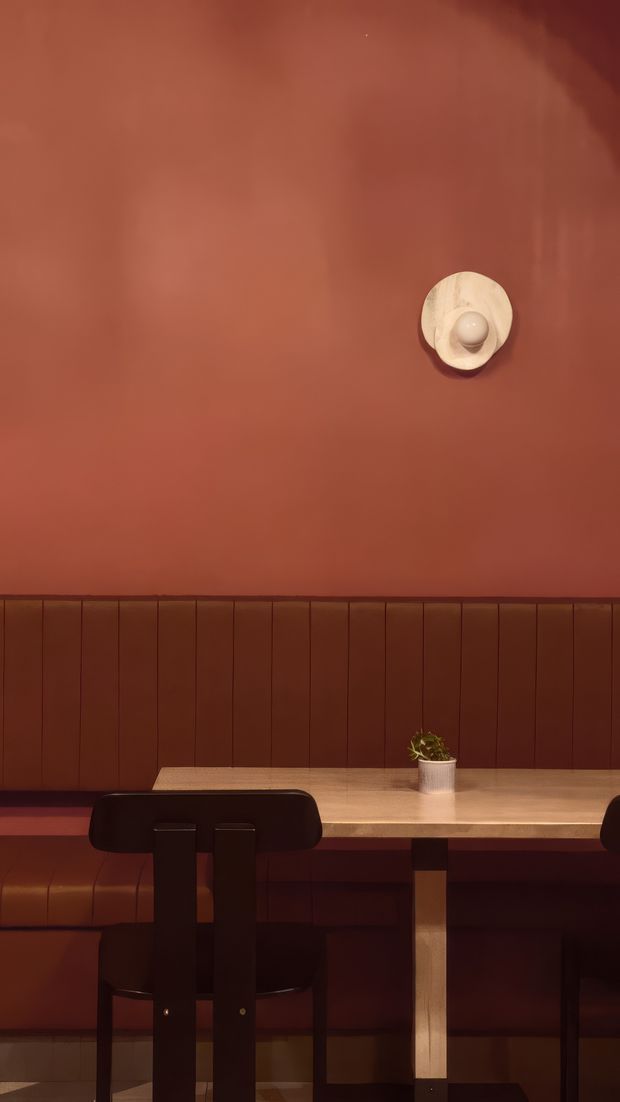 A moody, warm-toned dining nook with a terracotta-red painted wall, matching vertical timber wall panelling and integrated bench seating. A light timber dining table is paired with dark wood chairs, while a small sculptural wall light and potted plant provide restrained decorative accents.