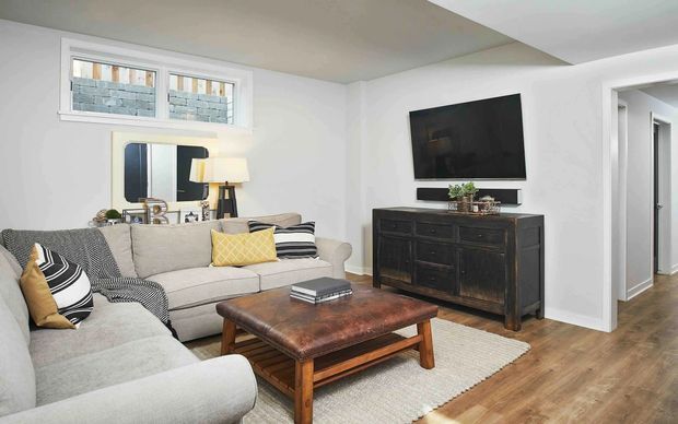 Bright, compact living room with a large L-shaped light fabric sectional, layered neutral cushions, a dark wood and leather-look coffee table, and a textured light rug. A dark timber sideboard supports a wall-mounted television, while a narrow console with decorative mirror and lamp sits beneath a small multi-pane window. White walls, simple recessed ceiling detailing, and warm timber flooring create a comfortable contemporary interior.