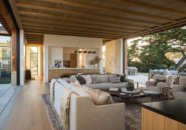 A spacious open-plan living room with a large sectional sofa, upholstered armchairs, a low wood coffee table and layered rug. The room opens to an outdoor terrace through wide glazing, with a kitchen and island visible beyond. Exposed timber ceiling beams and warm timber flooring contrast with light plaster walls, while a sculptural pendant and neutral furnishings create a refined, relaxed atmosphere.