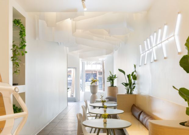 Bright, narrow contemporary café or hospitality interior with a predominantly white palette. The space features a row of small round tables and light chairs, a built-in tan upholstered bench along the right wall, abundant potted greenery, large glazed doors/windows at the rear, and distinctive sculptural white ceiling forms with linear wall lighting. A pale tiled floor and curved timber-toned joinery add warmth.