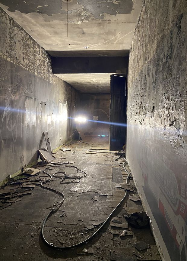 A narrow, dimly lit interior corridor under demolition or renovation. The space has heavily distressed concrete or plaster walls and ceiling, exposed wiring and cables, damaged flooring, construction debris, temporary work lights, and a dark doorway or opening at the far end. The raw, unfinished condition suggests an industrial or adaptive-reuse construction site.