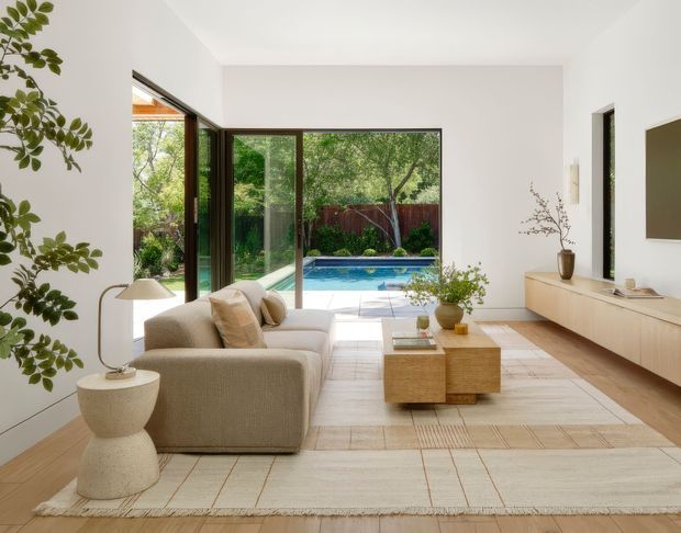 Bright, refined contemporary living room with a large beige sectional sofa, sculptural side table, low timber coffee table, and floating timber media cabinetry. Full-height glazed doors open directly to a landscaped garden and swimming pool, bringing abundant greenery and daylight into the room. The space has white walls, pale timber flooring, a large neutral rug, simple ceramics, and restrained natural styling.