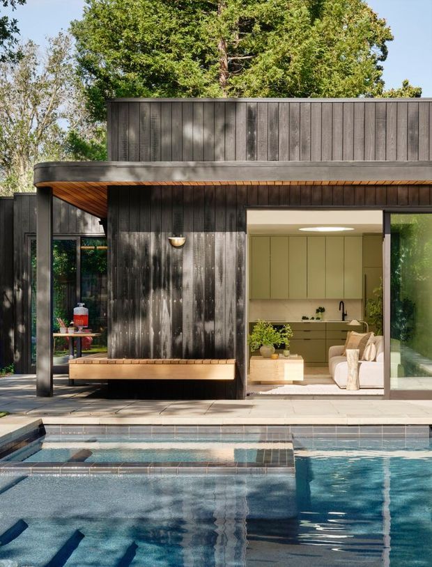 Exterior view of a modern, flat-roofed poolside house set among mature trees. The facade combines dark vertical timber cladding with broad glazing and a projecting timber-lined eave. A built-in bench sits beneath the cladding, while the open interior reveals a pale kitchen and living area. The foreground is dominated by a tiled swimming pool and surrounding paved terrace.