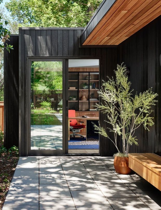 A contemporary black-clad courtyard entrance with a glazed door opening into a compact home office. The exterior features dark vertical timber cladding, a warm timber-lined projecting soffit, pale square paving, and integrated timber bench seating. Lush garden planting is visible around the entry, while the office interior includes built-in shelving and a red task chair.