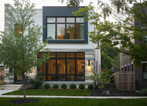 A contemporary multi-storey townhouse facade viewed from the street, with a strong geometric composition of dark-framed glazing, pale horizontal cladding, and grey masonry accents. The lower level has expansive windows revealing a warmly lit interior, while landscaped planting, a paved path, and timber fencing frame the frontage.