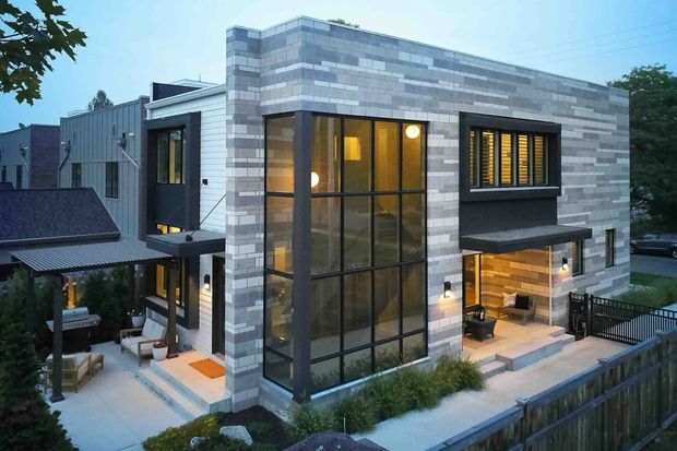 Contemporary multi-level residential house viewed from the exterior at dusk. The main facade features a projecting rectangular volume clad in horizontal bands of light and medium grey masonry, large dark-framed floor-to-ceiling glazing, and smaller dark-framed windows with exterior louvres or blinds. Warm interior lighting is visible through the glazing. A covered outdoor terrace and lower-level seating area sit beside landscaped planting, with a neighbouring roofline and timber boundary fence in the foreground.