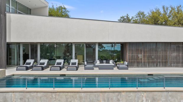 Contemporary multi-level residence viewed from the pool, featuring a long minimalist concrete or rendered facade, extensive floor-to-ceiling glazing, and a vertical timber-look privacy screen. A linear pool with stainless steel balustrade occupies the foreground, while a paved terrace is furnished with outdoor loungers and a compact seating arrangement.