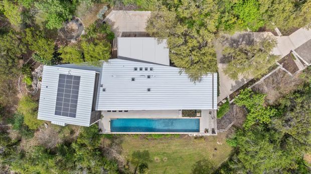 Aerial top-down view of a contemporary single-storey home set within dense landscaped vegetation. The building has a light metal roof with a large rectangular solar-panel array, adjoining roof forms, paved paths and terraces, and a long narrow turquoise swimming pool along the lower edge of the house.