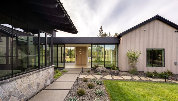 A contemporary single-storey home arranged around a landscaped courtyard. The composition shows a dark-framed glazed wing at left, a central glass-lined entry connector, and a pale vertical-clad gabled volume at right. Concrete stepping slabs, gravel planting beds, low shrubs, a small tree, and lawn create a structured garden approach. The architecture features strong black window and door frames, extensive glazing, a projecting dark roof, and a natural stone-clad base.