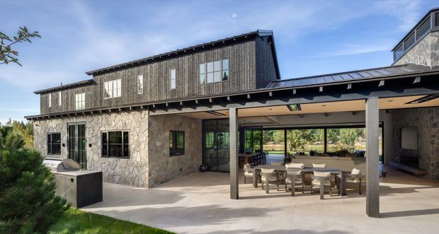 Exterior view of a substantial contemporary residence with a two-storey wing clad in weathered vertical timber boards above rough natural-stone walls. A covered outdoor dining and living area opens onto the landscaped grounds, with dark structural posts, a low-pitched metal roof, large glazed doors and windows, and a dining table beneath the covered terrace.