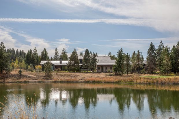 A low, single-storey rural home set among mature trees beside a calm pond or small lake. The building has a long, simple form, a muted grey corrugated pitched roof, and a restrained neutral facade partially screened by vegetation. The scene is photographed from across the water under a bright, lightly clouded sky, creating a quiet natural setting.