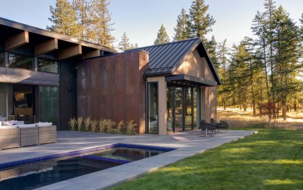 Exterior view of a modern dark-clad residence in a wooded setting, featuring a gabled metal-roofed wing, extensive floor-to-ceiling glazing, timber-toned wall cladding, a paved pool terrace, and a rectangular swimming pool. Outdoor lounge furniture sits beside the pool, with mature conifers and deciduous trees surrounding the property.