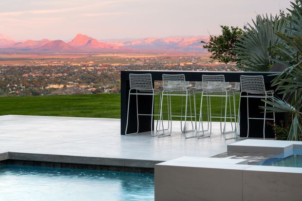 A contemporary outdoor entertaining area overlooking a broad valley and mountain range at sunset. The scene features a pale paved terrace, a swimming pool in the foreground, a raised black bar or outdoor counter with white wire bar stools, geometric built-in concrete or rendered pool elements, and sculptural drought-tolerant planting at the edge.