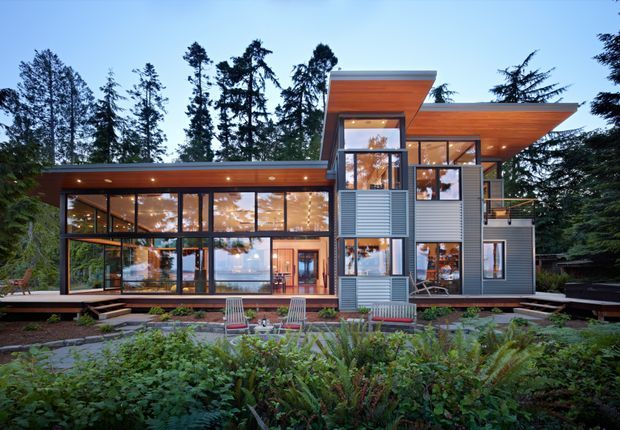 A contemporary multi-level residence set within a mature forested landscape. The facade combines extensive floor-to-ceiling glazing, dark window framing, grey horizontal cladding, and projecting flat roof planes with warm timber soffits. Outdoor seating and planted garden beds occupy the foreground, while the glass walls reveal illuminated interior spaces.