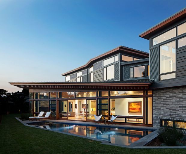 Exterior view of a large contemporary multi-level residence at dusk, with dark horizontal cladding, extensive floor-to-ceiling glazing, warm timber-toned window frames, and low-pitched roof forms. A covered outdoor entertaining area opens to a linear swimming pool and lawn, with the warmly lit living spaces visible through the glazing.