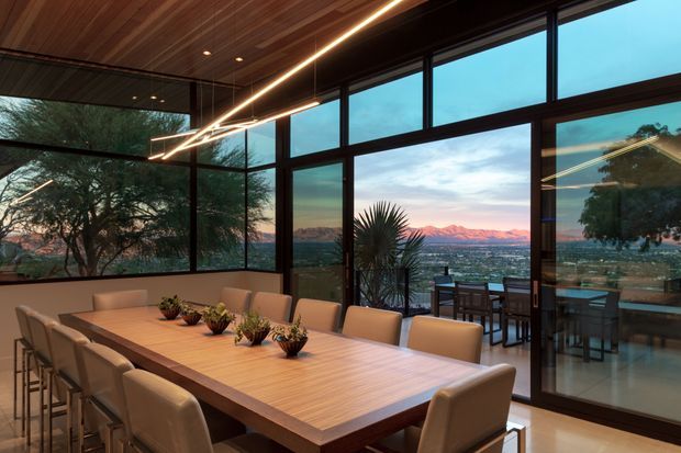 A contemporary dining area with a long timber dining table and upholstered chairs, positioned beside expansive black-framed floor-to-ceiling glazing. A timber-lined ceiling, linear pendant lighting, and warm interior finishes frame broad views toward a landscaped valley and distant mountains. An outdoor dining area is visible beyond the glass.