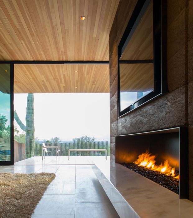 A contemporary indoor-outdoor living space with a large open glazed wall leading to a terrace and landscaped exterior. The room features a warm timber-lined ceiling with recessed spotlights, dark metal-framed glazing, pale large-format tiled flooring, a textured dark feature wall, and a long built-in linear fireplace beneath a reflective wall panel. Outdoor furniture, a tall cactus, and desert-like planting are visible beyond the opening.