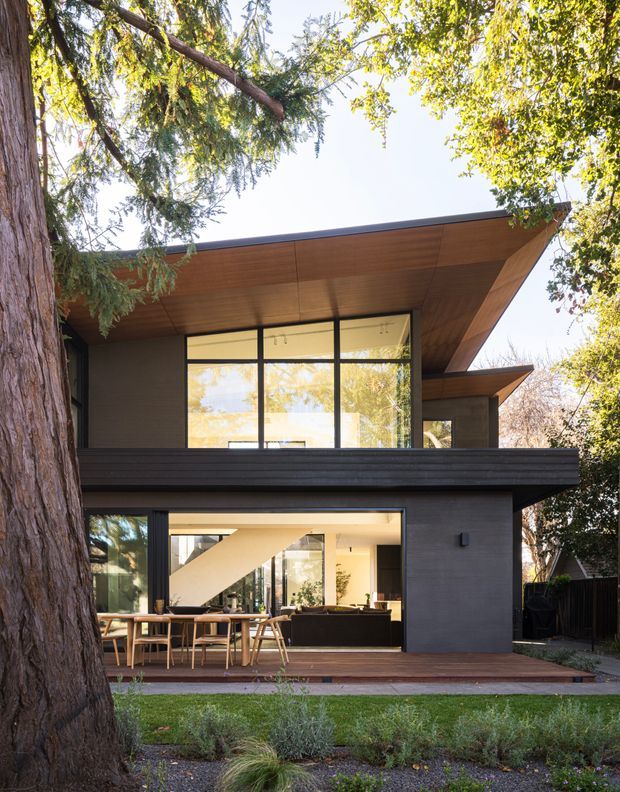 Exterior view of a contemporary two-storey home set among mature trees. The house features dark horizontal cladding, extensive black-framed glazing, broad cantilevered roof planes, and warm timber soffits. A timber deck and outdoor dining area connect to the glazed living spaces, with a visible internal stair and furniture beyond the windows.