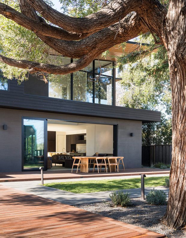 Exterior view of a contemporary two-storey residence set among mature trees. The dark horizontal-clad facade has extensive black-framed glazing, a projecting upper volume, and large open sliding doors connecting the kitchen and dining area to a timber deck. The landscaped foreground includes timber decking, lawn, gravel planting beds, and an established tree canopy.