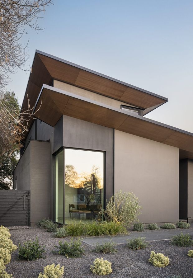 Low-angle exterior photograph of a contemporary, angular single-storey house. The facade combines smooth light rendered walls, dark charcoal panels, extensive warm timber soffits, and black-framed glazing. A projecting asymmetrical roofline and deep cantilevered eaves create a strong geometric composition. The home is set within a minimalist gravel landscape with low drought-tolerant planting.