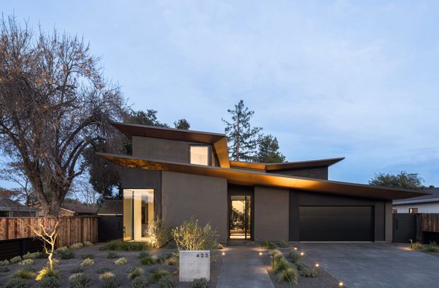 A contemporary single-storey residence viewed from the street at dusk, featuring a low-slung asymmetrical roofline with broad projecting eaves, dark rendered walls, warm illuminated glazing, and an integrated double garage. The front landscape uses gravel, drought-tolerant grasses, small shrubs, and architectural paving, with timber fencing enclosing the property.