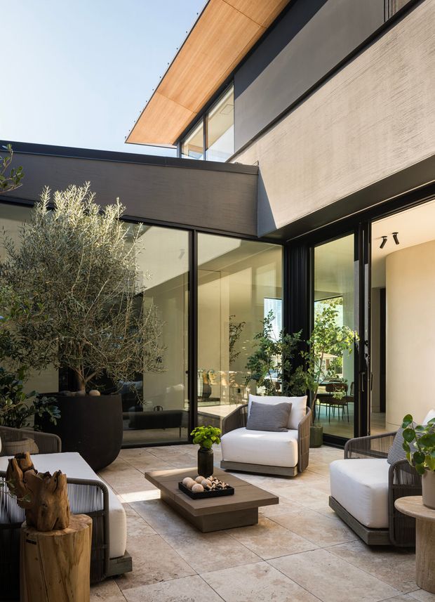 A sophisticated contemporary courtyard patio framed by dark cladding, expansive black-framed glazing, and a warm timber-lined roof overhang. The paved outdoor lounge contains white upholstered armchairs, woven side chairs, a low square coffee table, potted olive and leafy plants, and a sculptural timber stump. The courtyard opens visually into bright interior living spaces through the glazing and doors.