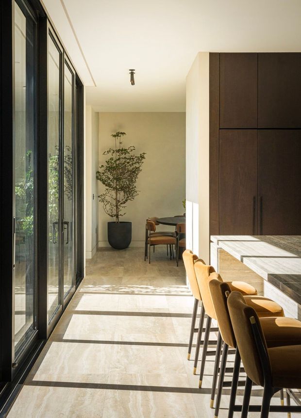 A refined open-plan interior with a long kitchen island and upholstered bar stools in the foreground, opening toward a dining area with a round table and chairs. Full-height black-framed glazing runs along the left side, casting strong linear sunlight across the pale stone floor. Dark timber cabinetry forms a tall kitchen wall on the right, with warm neutral walls, restrained detailing, and a large potted tree adding greenery.