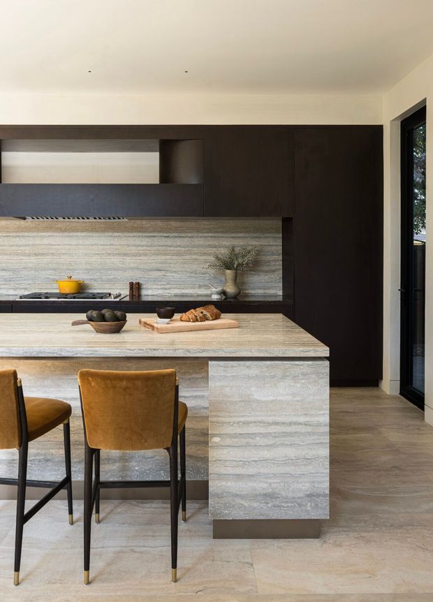 A refined contemporary kitchen featuring a large stone island with an integrated dining overhang and two ochre upholstered stools. Dark timber cabinetry spans the rear wall, contrasting with a horizontally veined stone splashback and matching island. A cooktop and rangehood are partially visible, with a tall dark pantry or storage wall beside black-framed glazing. The space is bright, minimal, and materially focused.