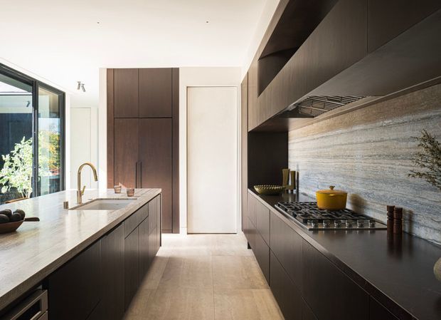 Contemporary galley-style kitchen with dark timber cabinetry, a large light stone island and benchtop, integrated appliances, and a textured horizontal stone splashback. Bright natural light enters through expansive glazing on the left, creating a refined, high-contrast interior.