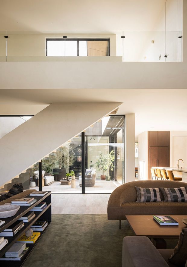 A refined contemporary open-plan living room with a dramatic sculptural staircase, double-height glazing, and a visible courtyard beyond. The space features a large curved brown sofa, low coffee table, dark open shelving filled with books, pale walls and floors, and a warm timber kitchen with island seating in the background. A glass balustrade overlooks the living area from the upper level.