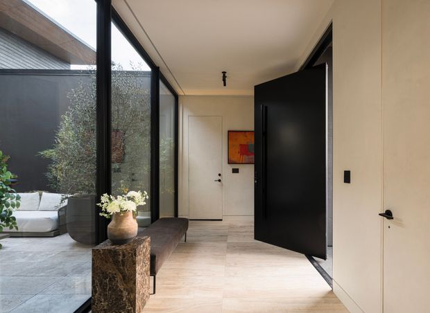 A refined contemporary entry hall with pale stone-look flooring, warm off-white walls, flush doors, and a dramatic oversized matte-black pivot door. Full-height black-framed glazing opens to a planted courtyard with outdoor seating, while a slim bench and stone pedestal with a vase add understated furnishings. A small abstract artwork provides a warm accent at the end of the hall.