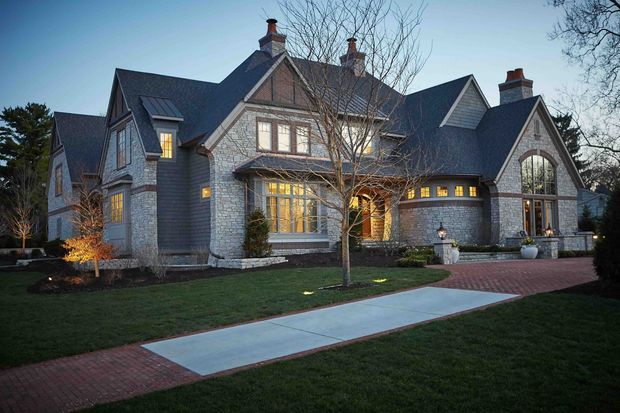 Large multi-gabled brick-and-shingle residence photographed at dusk, with steep slate-coloured pitched roofs, multiple brick chimneys, prominent arched window, projecting bay windows, and warm illumination from the windows and entry. The landscaped front garden includes lawns, a paved walkway, and a red brick driveway or forecourt.