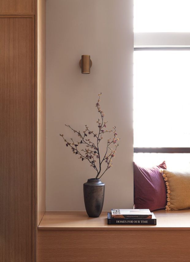 A warm, detail-focused interior vignette showing a built-in timber window seat and cabinetry, a bronze wall sconce, a dark ceramic vase with branches, and stacked books. The window seat includes burgundy and mustard cushions, with bright daylight entering through the large window. The composition has a refined, contemporary feel with natural wood and muted earthy tones.