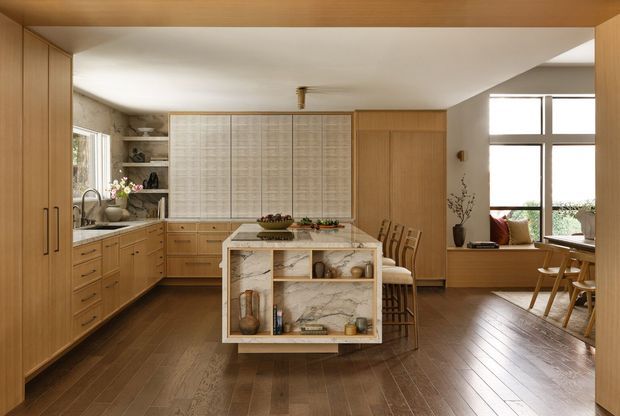 Warm, open-plan kitchen and dining area with extensive light timber cabinetry, a marble-featured island with open shelving, integrated shelving and a large corner window. The kitchen opens directly into a bright dining or living zone with timber flooring and mid-century-inspired furnishings.