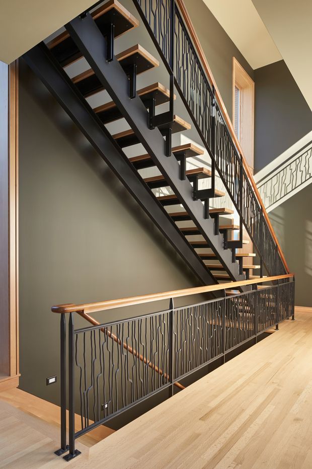 Interior view of a modern stairway and upper-level landing. An open-riser staircase with black steel stringers and supports, light timber treads, and distinctive vertically wavy black metal balustrades rises through a tall stair void. Warm timber handrails and pale timber flooring contrast with dark grey walls and black metalwork, while a tall timber-framed window brings in natural light.