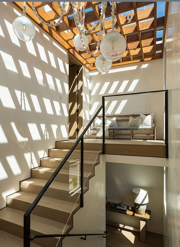 Bright contemporary interior stairwell and landing with a timber staircase, black metal and glass balustrade, exposed timber roof beams with translucent skylight panels, and multiple sculptural glass pendant lights. The upper landing contains a built-in or compact timber-framed daybed with cushions, while a small console and lamp are visible below. Strong geometric sunlight patterns fall across the pale walls and timber surfaces.