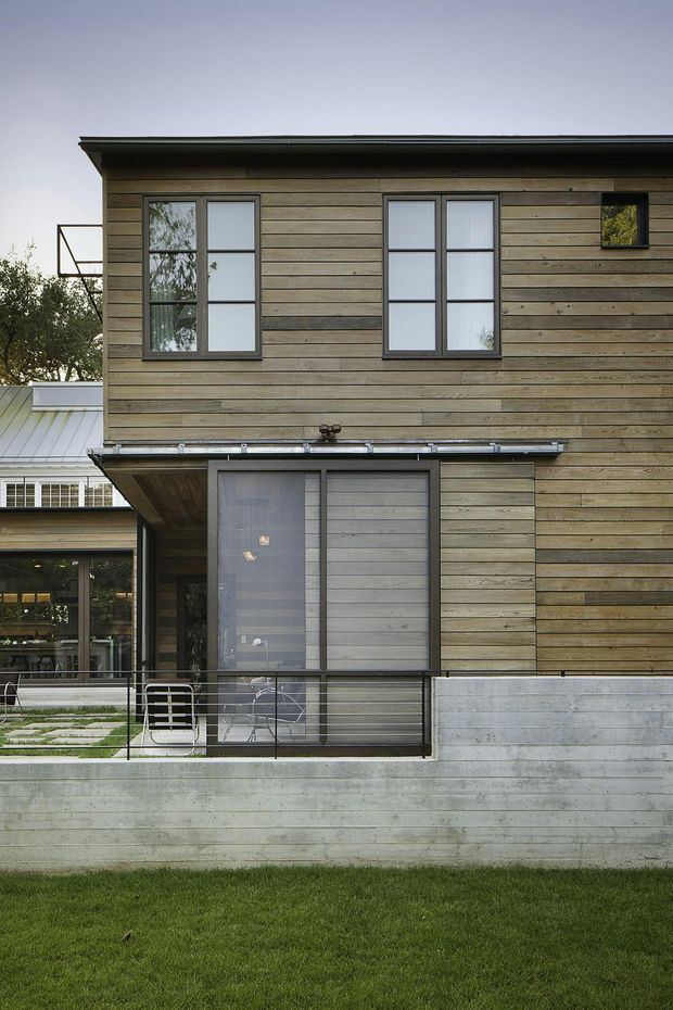 Exterior view of a contemporary two-storey house with horizontal timber cladding, dark-framed rectangular windows, a low-pitched roof, and a concrete lower level. A covered terrace or balcony with simple metal railings sits above a lawn, while glazed living areas and a lighter adjoining structure are visible to the left.