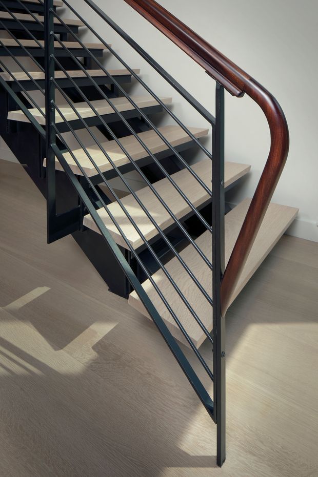 Close-up architectural photograph of a contemporary open-riser staircase. Pale timber treads are supported by a dark metal stringer and framed by dark metal balustrades with horizontal rods. A prominent curved, polished dark timber handrail runs along the stair edge. The staircase is set against a light neutral wall and timber flooring, with strong sunlight creating shadows across the floor.