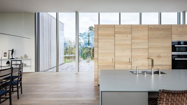 A bright, open-plan contemporary kitchen and dining area with a large pale grey island, integrated sink and tapware, full-height light timber cabinetry, and built-in black appliances. Light timber flooring extends toward full-height glazing and an outdoor deck, while a simple dining table and shelving sit to the left.