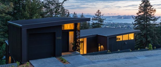 Contemporary dark-clad hillside residence viewed from the street at dusk. The low-slung composition includes a prominent garage volume and connected living volumes with flat or very low-pitched roofs, vertical cladding, and warm illuminated glazing. A bay and surrounding tree-covered hillside are visible beyond the home.