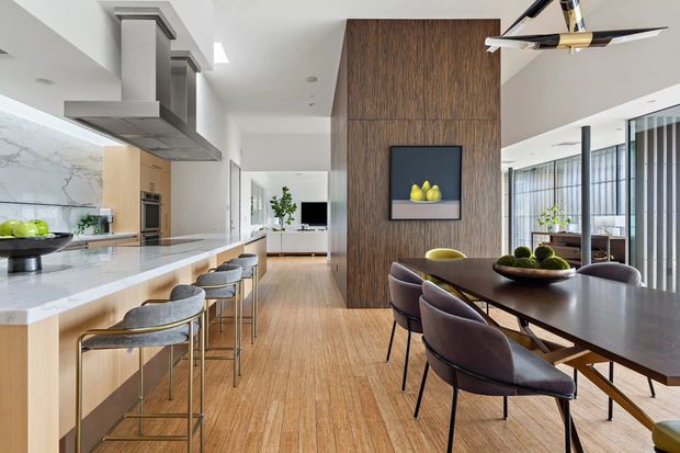 Open-plan contemporary kitchen and dining area with a long pale stone island, integrated stainless-steel appliances, sculptural rangehood, timber cabinetry, and several bar stools. The dining zone contains a dark timber table with upholstered chairs, set against a tall dark timber feature wall with artwork. Full-height glazing with vertical screening brings in abundant natural light, and light timber flooring extends throughout.
