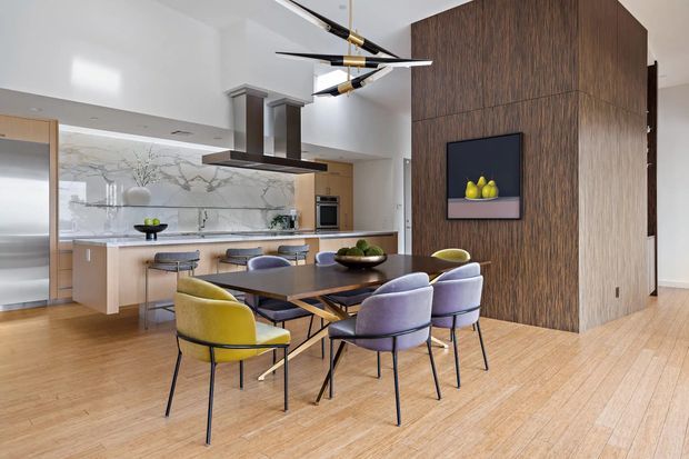 Open-plan contemporary kitchen and dining area with a warm timber floor, pale wood cabinetry, a large marble-look splashback, and a central island with integrated cooktop and sculptural rangehood. A dark vertical timber feature wall separates the kitchen from the dining zone and displays artwork. The dining table is surrounded by colourful upholstered chairs, while a linear modern pendant hangs overhead.