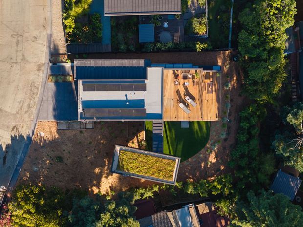Top-down aerial view of a contemporary residential property in a leafy setting. The house has a long rectangular footprint with a flat or low-pitched roof, a large timber outdoor terrace with lounge furniture, and a separate rectangular green-roofed structure in the yard. Dense mature planting surrounds the site, with a paved street along the left edge.