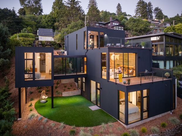A multi-level contemporary home on a steep, wooded suburban site. The building is composed of dark charcoal-clad geometric volumes with extensive floor-to-ceiling glazing, projecting balconies, and connecting glazed sections. Warm interior lighting is visible through the windows, while landscaped terraces, a small lawn, and planted garden beds surround the house.