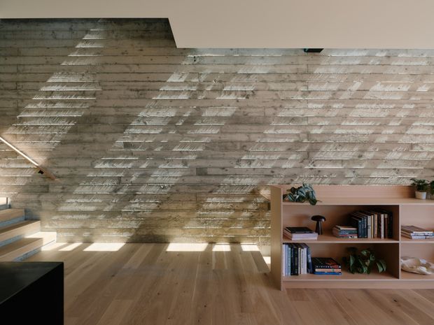 Interior view of a contemporary living space with a full-height textured concrete feature wall, warm timber flooring, and built-in timber shelving filled with books and plants. Strong patterned sunlight filters across the wall and floor, while a partial staircase and dark furniture are visible at the edges of the frame.