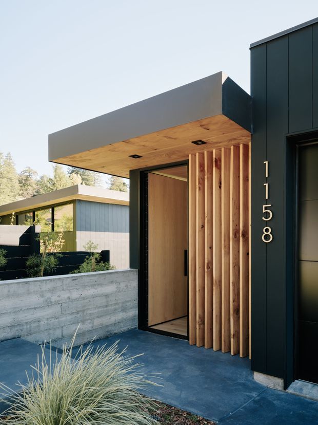 A modern residential entry facade viewed from the front walkway. The design features dark vertical cladding, a projecting flat canopy with a warm timber soffit, a large timber pivot door, and a vertical timber slat screen. Concrete paving and a low raw concrete planter wall frame the entrance, with a neighboring modern wing visible in the background.