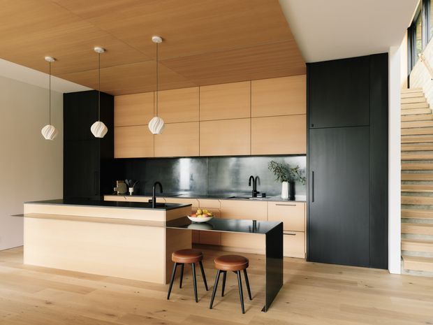 Contemporary open-plan kitchen with extensive light timber cabinetry and matching timber ceiling, matte black full-height joinery, a black stone island and breakfast bar, and a dark metallic splashback. Three pendant lights hang above the main island, while a staircase is visible at right. The space is clean, warm, and refined with strong black-and-timber contrast.