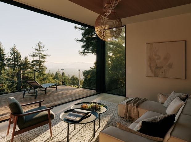 A warm, refined contemporary living room opening onto a timber deck through expansive corner glazing. The room contains a large upholstered sofa, sculptural lounge chairs, nested round coffee tables, a textured area rug, abstract wall art and a woven pendant light. Tall trees and a distant landscape are visible beyond the deck, creating a strong indoor–outdoor connection.